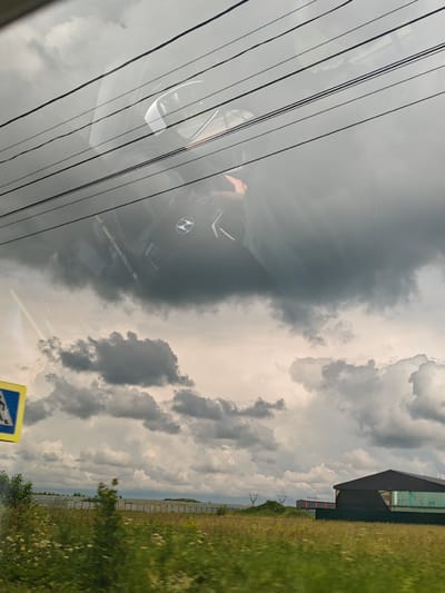 The image captures a view from inside a vehicle, looking out through the windshield. Overhead, multiple dark power lines crisscross the frame. The sky is filled with dramatic, dark, and light gray clouds, suggesting overcast or stormy weather.
Through the windshield and partially obscured by the power lines and clouds, a reflection of the vehicle's interior is visible. This reflection shows a steering wheel with the Hyundai logo in the center, along with parts of the dashboard and what appears to be a person's head and shoulder.
In the lower portion of the image, a green field with various plants and wildflowers stretches towards a fence line. Beyond the fence, there are some structures, including a dark-roofed building with green siding and what appears to be a smaller building or shipping containers. There are also some industrial-looking towers or pylons in the distance. A yellow and blue pedestrian crossing sign is visible on the left side of the frame. The scene is set in or near Repnikovo, Russia.