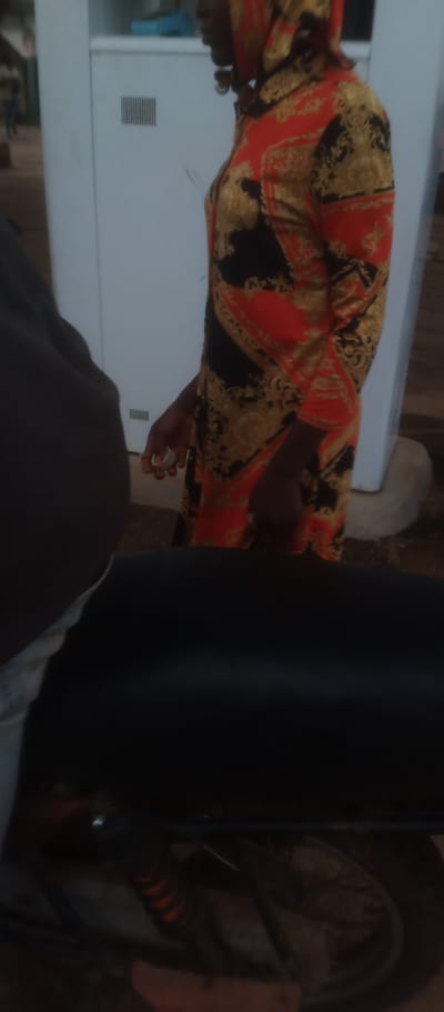 The image shows a side profile of a person wearing a hooded garment with a vibrant red, black, and gold pattern. The garment appears to be a long shirt or dress, with a hood pulled up over their head. The person's hands are visible; one hand is holding a small, round white object. The background consists of a pale, light-colored structure, possibly a building or a large appliance, with a rectangular vent on its surface. To the left of the person, a dark, out-of-focus object is partially visible, which might be another person or an item of clothing. The lower portion of the image is dominated by a dark, out-of-focus area that appears to be a motorcycle or bicycle seat and wheel, suggesting the scene is outdoors. The ground appears to be dirt or gravel. This scene is located in Kukum, Nigeria.