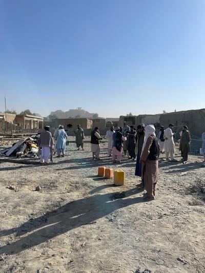 The image depicts an outdoor scene in Spin Boldak, Afghanistan, under a clear, bright blue sky. Approximately 15 to 20 adult males, dressed in traditional Afghan clothing consisting of shalwar kameez, vests, and head coverings, are gathered on uneven, dusty ground. They are observed standing in small groups, appearing to be conversing or observing their surroundings. Long shadows extend from the individuals across the ground.
In the foreground, three plastic jerrycans (two orange and one yellow) are positioned on the dirt. To the left mid-ground, a disorganized pile of debris is visible, comprising corrugated metal sheets, wooden planks, and miscellaneous discarded items, with a white plastic chair positioned nearby. The background features a desolate landscape of numerous damaged or partially constructed mud-brick buildings and walls. In the distant background, a more prominent, fortified-looking structure or settlement is discernible on an elevated terrain, partially obscured by atmospheric haze. There are no direct interactions such as handshakes or specific actions beyond standing and grouping.