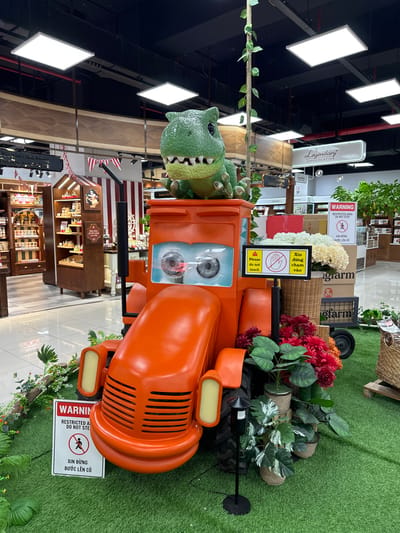 This image shows a large, orange, cartoon-like tractor with a green dinosaur figure sitting on top. The dinosaur has large eyes and an open mouth with sharp teeth. The tractor has a black exhaust pipe and large, round headlights. Around the base of the tractor, there are artificial plants, flowers, and a small black lamp post. There are also signs with warnings in English and Vietnamese. One sign reads "WARNING RESTRICTED AREA DO NOT STEP". Another sign reads "Please do not touch".
In the background, there are shelves filled with products, a large display of flowers, and signs that say "Legendary Chocolatier". The scene appears to be set up in a commercial establishment, possibly a department store or a supermarket, with a polished floor and overhead lighting. The location is identified as Phu Thuy, Vietnam.