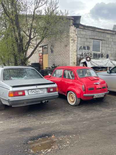 The image shows a street scene in Mar’ino, Russia. A red compact car and a silver BMW 7-Series are parked next to each other. Behind the cars is a weathered, two-story brick building with graffiti and a pile of tires. A tree with fresh green leaves is visible on the left. The street is paved with asphalt and shows signs of disrepair, including potholes. The sky is overcast. A sign on the back of the silver car reads: 'T542BO 799'. The license plate on the red car appears to read: 'M739HM 199'.