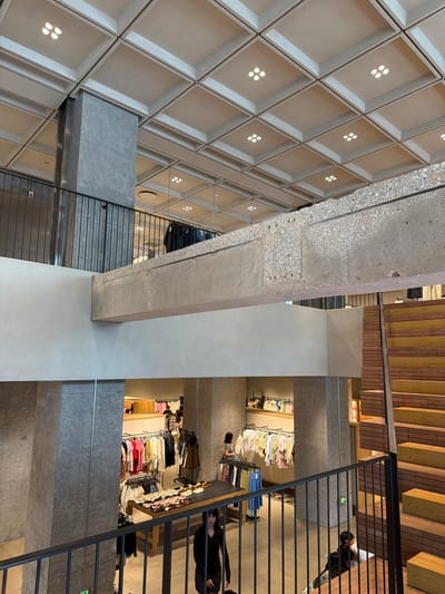 The image shows the interior of a multi-level retail store, possibly a clothing boutique, located in Qinhuai District, China. The architectural style is modern industrial, with exposed concrete columns and beams, a coffered ceiling with recessed lighting, and a prominent staircase made of wood with a striking orange and yellow hue.

On the upper level, a black metal railing borders a balcony overlooking the lower level. Clothing racks filled with various garments, including shirts and dresses in a range of colors, are visible. A person, dressed in dark clothing, is standing near the railing.

The lower level features more clothing displays, including racks of shirts and a table showcasing shoes. Several individuals are present: one person in a black outfit is looking down, another person, partially visible, is walking, and two individuals are seated on the wooden stairs. The environment appears to be a well-lit, organized retail space with a focus on fashion merchandise.
