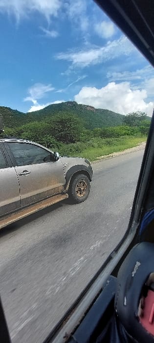 A forensic examination of the image reveals a dynamic scene captured from within a moving vehicle in Ruaha Darajani, Tanzania. The primary subject is a silver/grey pickup truck, heavily coated in dirt and mud, traveling on a paved asphalt road. This truck exhibits black fender flares, bronze-colored multi-spoke wheels fitted with off-road tires, and visible side steps also covered in debris. A partial roof rack is discernible above the front door.

The background consists of verdant, densely vegetated hills and mountains with scattered rocky outcrops, extending towards a bright blue sky marked by sparse white clouds. The foreground includes elements of the observer's vehicle: a dark window frame and pillar are visible on the right side of the frame, and a dark blue object with a red accent, consistent with a vehicle's interior component, is partially visible in the lower right corner. Both vehicles appear to be in motion, traveling in the same general direction. No individuals are identifiable within the frame.