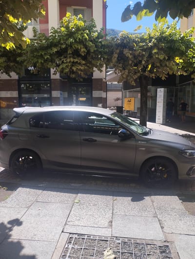 The image depicts a sunlit urban street scene in Bellinzona, Switzerland. A dark grey, possibly matte-finished, Fiat Tipo hatchback is parked prominently on a stone-tiled sidewalk, facing right. The car features "SPORT" badging on its fender, black multi-spoke wheels with visible yellow brake calipers, and a rear spoiler.
Overhead, trees with lush green leaves provide shade, casting distinct shadows on the sidewalk and parts of the car. In the background, several multi-story buildings line the street. One building has a reddish facade with white window shutters and balconies, while others are lighter in color with glass storefronts. Two men are visible near a shop entrance on the right side of the image, appearing to walk or stand. Majestic mountains rise in the far background under a clear blue sky, indicating the mountainous terrain of the region.
The scene takes place during the daytime under clear, sunny weather, as evidenced by the bright light and strong shadows.
Visible text includes "SPORT" on the car. A yellow sign displays "AGG" and "PHOTO GALLERY & FRAMES". A tall, white vertical sign or pillar clearly shows "32 Via V. D'Albono", confirming the street and location. This sign also lists "2.5L", "0.5L", and "BOUQUET" below the address, likely product information or a directory. Storefront windows show "SPACE" and a "JM" logo.