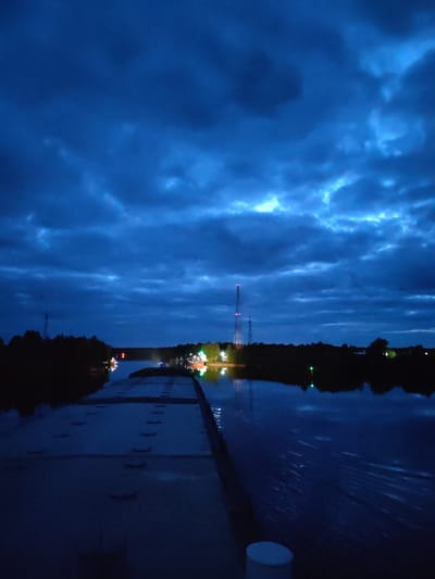The image depicts a nighttime scene from Annensky Most, Russia. A concrete structure stretches into a dark body of water. Reflections of lights are visible on the water's surface. On the opposite shore, silhouetted trees line the horizon, with a tall communication tower rising above them. The sky is filled with heavy, dark clouds. The overall lighting suggests a low-light or dusk setting.