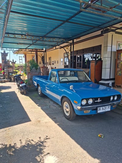 The image shows a blue Datsun pickup truck parked on a paved street in Lodtunduh, Indonesia. The license plate number is visible as DK 8439 PT. Next to the truck is a parked motorcycle. The street is lined with buildings featuring Balinese architectural elements, including ornate carvings and tiered structures. There is a covered walkway with a corrugated metal roof alongside the road. In the foreground, there are flowers and offerings on the road. The lighting indicates it is daytime.
