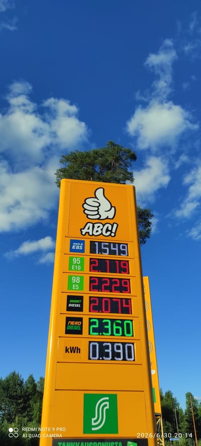 This is a media file captured from a low angle, looking up at a bright yellow gas station sign against a vibrant blue sky filled with fluffy white clouds. The sun is shining, indicating it's daytime. The sign displays prices for different fuel types and electricity.
The prominent subjects are the fuel price displays on the sign. The prices shown are: EKO E85 at 1.549, 95 E10 at 2.119, 98 E5 at 2.229, SMART DIESEL at 2.074, NERO DIESEL at 2.360, and kWh at 0.390. The gas station's logo, a white hand pointing with "ABC!" written next to it, is also visible. Additionally, a green logo with a stylized "S" is displayed on the right side of the sign, and text reading "TANKKAUSBONUSTA" is present.
The scene is set at a gas station, likely in Joutsa, Finland, given the text on the sign. The weather appears to be clear and sunny with scattered clouds. A timestamp in the upper right corner of the image indicates "2026/6/30 20:14," although this might be a misinterpretation or a future date.
A redmi note 9 pro AI quad camera watermark is visible at the bottom left corner, suggesting the image was taken with this device. The overall impression is a clear, sunny day at a Finnish gas station, with the focus on the fuel prices and the branding of the establishment.