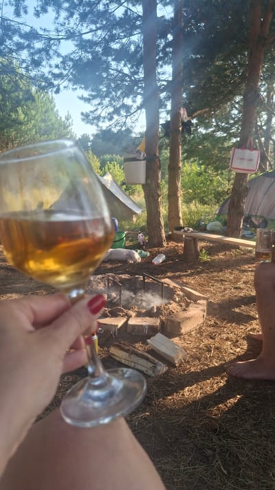 A hand with red nail polish holds a wine glass filled with amber liquid in the foreground, obscuring part of a campsite. In the mid-ground, a campfire pit made of bricks is smoking gently, with logs and debris scattered around it. A white and pink tote bag with the text "CVMARTANNI" is hanging from a tree. Various camping tents are visible in the background, along with trees and foliage, suggesting an outdoor setting. The ground is covered with pine needles and dry grass, indicative of a forest environment. Sunlight filters through the trees, creating dappled shadows. The scene appears to be set in the Навасвержанскі сельскі Савет region of Belarus.