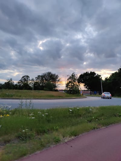 The image captures an outdoor scene at either sunrise or sunset, centered around a roundabout. The sky is largely covered by dense, grey and purplish clouds, but streaks of orange and pink light peek through at the horizon, particularly behind the trees on the right and through breaks in the clouds, casting a dramatic glow.
A silver or light-colored hatchback car is visible on the right side of the roundabout, its rear lights illuminated, suggesting it is braking or has stopped. The car features a yellow license plate. The roundabout's central island is covered in green grass, with some patches of white and yellow wildflowers. Similar grassy verges line the road, and a reddish asphalt path, likely a bicycle lane, occupies the foreground on the right.
Along the distant horizon, a line of dark, leafy trees stands against the lighter sky. Several streetlights are also present along the roadside. The overall setting appears to be a suburban or semi-rural area, characterized by road infrastructure, natural greenery, and overhead clouds. No people are visible in the frame. The scene is set in Oosterhout, Netherlands.