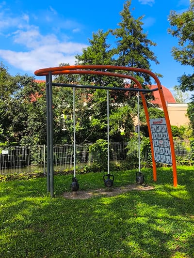An outdoor fitness station is centrally located within a grassy park area in Oberschaeffolsheim, France. The primary structure is a large, orange, curved metal arch with gray support poles. Three black kettlebells are suspended from the top bar by vertical silver poles, resting on circular worn dirt patches on the green grass. A vertical orange sign attached to the right pole displays "KOMPAN" at the top and multiple black-and-white exercise illustrations below. In the midground, a light-colored metal fence acts as a barrier along the edge of a narrow body of water, possibly a canal or stream. Dense green foliage, comprising trees and bushes, forms a thick backdrop behind the water and fence, with parts of buildings featuring reddish-orange tiled roofs visible through the distant greenery. The scene is illuminated by bright daylight under a clear blue sky with scattered white clouds. No individuals are present in the image.