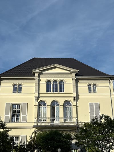 The image shows the facade of a light yellow, two-story building with a dark gray, tiled roof. The building has several windows, some with arched tops and white shutters. A balcony with a metal railing extends across the central portion of the upper floor. Trees with dark green foliage are visible in the foreground and partially obscure the lower level of the building. The sky above is a clear blue with a few wispy white clouds. The architectural style suggests it could be a historic building. This building is located on the Kaiserstraße in Bonn, Germany.