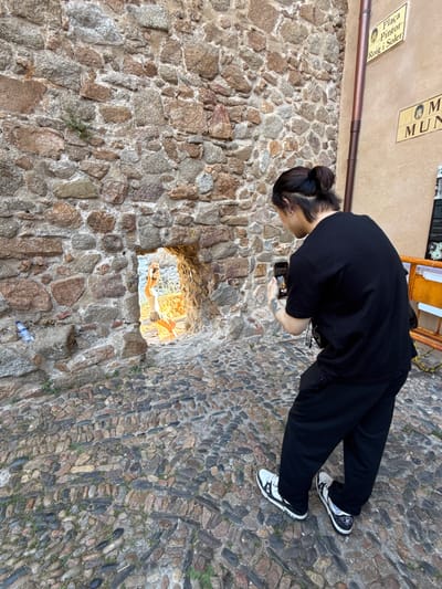 A person with dark hair pulled into a bun is photographing another person through a hole in a stone wall. The person being photographed is standing in what appears to be a narrow passageway illuminated by sunlight, wearing a light-colored top and a skirt. The photographer is wearing a black t-shirt and black pants, and holding a smartphone in their right hand. The wall is constructed of irregular stones of various colors, and the ground is paved with small, smooth stones. In the background, signs are visible on a painted wall, one reading "Placa Pintor Roig i Soler" and another with the word "MUN" visible. The scene is located in Tossa de Mar, Spain.