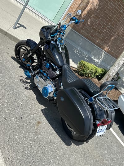 This media file features a customized black Harley-Davidson motorcycle parked outdoors on a paved surface, likely a parking lot, in Port Coquitlam, Canada. The motorcycle is the primary subject, adorned with numerous blue accents on parts such as the handlebars, footpegs, engine components, and mirrors, suggesting a personalized aesthetic. The seat has a diamond-stitched pattern. A chrome luggage rack is mounted on the rear of the motorcycle, above hard saddlebags. The motorcycle has a license plate visible with the text "4743A5" and "BRITISH COLUMBIA".

The scene appears to be set during the daytime, under bright sunlight, as indicated by the strong shadows cast by the motorcycle and a nearby tree. The weather seems to be fair and warm, contributing to the outdoor setting. In the background, there is a brick building and some green shrubbery. A portion of a white vehicle is also visible parked to the right of the motorcycle. The ground is asphalt, with some lighter colored concrete areas near the building. No people are visible in the frame, and the overall mood is static, focusing solely on the parked vehicle. The image is taken from a slightly elevated, angled perspective.