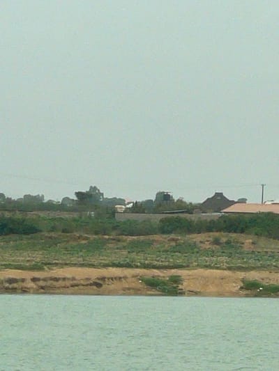 The image presents a view of the landscape adjacent to what is purported to be Nabatum, Nigeria. The scene includes a body of water to the right, with a shoreline comprised of vegetation, soil, and various unidentified materials. Beyond the immediate shoreline, there are indications of buildings or structures and trees situated further inland. The sky is overcast, creating a diffused ambient lighting condition. Due to the resolution and quality, specific details about the individual buildings, the nature of the vegetation, or the composition of the soil cannot be ascertained. There is an absence of discernible human activity or presence within the field of view.