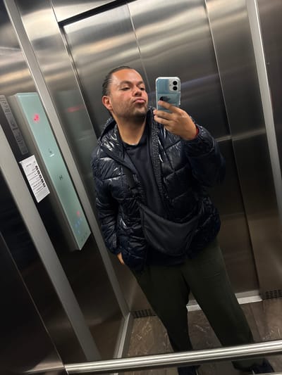 A man takes a selfie in an elevator, holding a cell phone in his right hand. He is wearing a dark puffer jacket and dark green pants. The elevator has stainless steel walls and a control panel with illuminated buttons. A small, white sign with black text is affixed near the control panel. The location of the photograph is inside the elevator in Tizapan, Mexico.