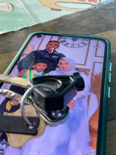 A smartphone with a green case displays a family portrait as the wallpaper. The wallpaper shows a man in a dark blue patterned shirt, a young boy in a graduation cap and gown in green and yellow, and a woman wearing a light blue hijab. The time on the phone screen reads 13:26, with the date "14 Jun". In the foreground, a set of keys is placed over the lower portion of the phone screen, obscuring part of the image and partially covering the keys. The background appears to be a wooden surface with a textured, patterned mat or cloth. The scene is set on a table, likely in an indoor environment. The location in Meurya, Indonesia, is not discernible from the visual information provided in the image.