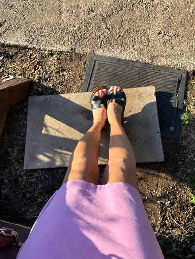 A first-person perspective shot captures a person's legs and feet from above, seated on a wooden bench. The person is wearing black sandals and a purple skirt. The ground is a mix of dirt, gravel, and wood chips. In the background, a concrete slab and a black rubber mat are visible. The scene is illuminated by bright sunlight, casting distinct shadows. The location appears to be an outdoor setting, potentially a backyard or a park area within Verkhneyarkeyevo, Russia.