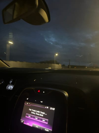 The image depicts the interior perspective from a moving vehicle on a road in São Mamede de Infesta, Portugal, at 17:47, under a dark, cloudy sky. The vehicle's dashboard is visible in the foreground, with a digital display indicating a speed of 124 KM/H on the left. Centrally, an infotainment screen displays "Fair Trade (with Travis ...)" by "Drake, Travis Scott" as the current song playing via Bluetooth, with playback controls and menu options ("Leitor," "Lista," "Opções") in Portuguese. A red hazard warning light button is visible above the screen. Through the windshield, a continuous light-colored barrier or wall borders the road on the left. Multiple tall streetlights with bright yellow illumination are present, contrasting with the dark, overcast background sky. The top of the vehicle's rearview mirror is visible at the top of the frame.