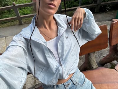 A person is seated at a wooden table in an outdoor setting, appearing to be located in Sà Phìn Commune, Vietnam. The individual is wearing blue denim shorts and a light blue zipper jacket over a white ribbed tank top. A gold necklace is visible around the neck. The table appears to be part of a rustic outdoor setting, possibly a park or garden, given the presence of greenery visible in the background through what seems to be a fence. The person's hand is visible near the jacket, and two dark braids frame the face. The photo is taken from a slightly angled, top-down perspective.