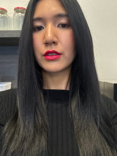 A female individual is depicted in a close-up, chest-up portrait. The subject has long, straight, dark hair, transitioning from black roots to lighter, possibly gray or dark brown, ends. She is wearing a black, ribbed, high-necked, long-sleeved top. Her face is directed towards the camera, featuring bright pink lipstick and visible eye makeup. In the background, behind the subject's head and to the upper left, a dark-colored shelf is visible, upon which two empty, clear glass jars with red lids are placed. A light-colored wall forms the background behind the shelf and extends to the right. To the lower left, a white translucent plastic container with a blue lid rim is partially visible on a surface. To the far right, a dark, rectangular object is partially visible. The specific location within Herentals, Belgium, cannot be determined from the visual information provided.