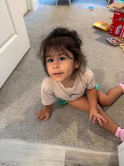 A young girl is lying on a grey carpeted floor. She is wearing a light beige t-shirt and a turquoise skirt. She has dark brown hair that is pulled back, and her eyes are looking up at the camera. Her left leg is bent, and her right leg is extended. Her right hand is reaching out, with her fingers splayed.
In the background, there are some objects on the carpet, including a red box with an image of a child on it, and a yellow and orange box. There is also a blue and orange toy on the floor. To the right of the carpet, there is a light-colored wooden floor. The image appears to be taken indoors during the daytime. The girl's expression is neutral, and she does not seem to be experiencing any strong emotions.
There is some text visible on the red box in the background. It appears to say "HUGGIES" and "LITTLE MOVERS". There is also a number "6" visible on the red box.