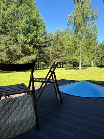 The image depicts an outdoor scene in Копнино, Russia, under clear, bright daylight. In the foreground, a dark gray or brown composite deck extends across the bottom, featuring horizontal planking. On the deck, two dark wooden folding chairs are partially visible. The chair on the left shows its backrest and part of its slatted seat, over which a light beige, quilted cushion with visible ties is draped. To the right, another similar wooden chair appears partially folded or angled. Further right on the deck, a light blue, conical or dome-shaped object, possibly a cover or part of an outdoor fixture, is visible. Beyond the deck, a vibrant, meticulously maintained green lawn spans the midground, illuminated by direct sunlight. The background consists of a dense treeline, featuring numerous tall pine trees with dark green foliage and several birches, identifiable by their light-colored bark and brighter green, delicate leaves. The sky above is a uniform, clear blue with no clouds. No individuals or animals are present in the scene.