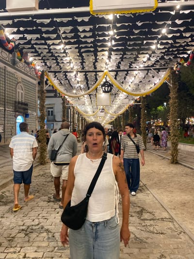 A medium shot shows a woman in the foreground with her mouth open, facing the camera. She is wearing a white tank top and blue jeans, with a black crossbody bag. Behind her, a cobblestone street is lined with people walking in both directions. The street is decorated with strings of white paper flags and yellow and white fabric garlands, suspended from lampposts and overhead structures. In the background, buildings with patterned tile facades are visible. The scene appears to be a street festival or celebration in Tavira, Portugal.