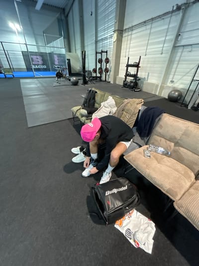 In a gym setting, a man wearing a pink hat sits on a couch, tying his shoelaces. A black Bullpadel backpack rests next to him. Various gym equipment, including weight racks and a balance ball, are visible in the background. A water bottle and a phone are on the couch near the man.