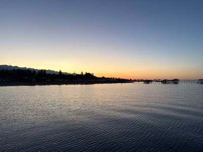 The image depicts a serene sunset over Issyk-Kul Lake in Sary-Oy, Kyrgyzstan. The sky transitions from a deep blue at the top to a warm yellow and orange glow near the horizon, casting a soft light over the water. In the distance, a silhouetted mountain range with sparse vegetation lines the shore. A beach with scattered umbrellas and structures is visible on the left side of the frame. On the right, a long pier extends into the lake, with several small, enclosed structures perched on stilts, suggesting a resort or recreational area. The water itself is calm, with gentle ripples reflecting the colors of the sky. There are no people visible in the scene. The overall impression is one of tranquility and natural beauty at the end of the day.