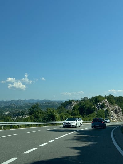 The image captures a view from a vehicle traveling along a multi-lane highway through a mountainous, treed landscape. The highway features white lane markings and a metal guardrail on the left. Several cars are visible on the road; a white SUV is in the left lane, a dark blue sedan is in the right lane, and another car is further ahead in the right lane. The background consists of rolling hills covered in dense green vegetation, with more distant, rugged mountains under a clear blue sky with scattered wispy clouds. The scene appears to be a scenic route, possibly in a rural or semi-rural area. The location is identified as Očinići, Montenegro.