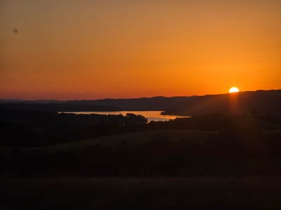 The image depicts a serene sunset over rolling hills and a body of water in Golyama Zhelyazna, Bulgaria. The sun, a bright orange orb, is partially obscured by a distant hilltop, casting a warm, golden glow across the sky. The sky transitions from a deep orange near the horizon to a softer, lighter orange at the top. Silhouetted against this vibrant sky are layers of dark, undulating hills, suggesting a rural landscape. A body of water, possibly a lake or river, is visible in the middle ground, reflecting the sunset's colors. In the foreground, the land is dark and indistinct, with hints of grass or foliage. The overall impression is one of tranquility and natural beauty, captured during the twilight hours.