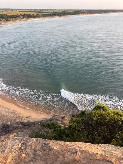This media file is an aerial, vertical shot of a beach and ocean scene. The image is oriented such that the ocean is on the left and the shore is on the right. The water is a deep teal color with subtle ripples, indicating calm conditions. A gentle wave is breaking on the shore, creating frothy white foam. The beach is sandy and appears to be bordered by lush green vegetation and rocky outcrops. In the distance, a stretch of land with green fields and scattered trees is visible, suggesting a rural or natural coastal environment. The lighting suggests it is late afternoon or early evening, with the sun casting warm tones on the rocky cliff on the far right. There is no visible text or people in the image. The location is identified as Shastrawela, Sri Lanka.