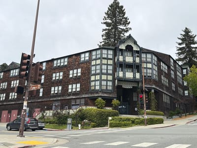 The image depicts an exterior view of the Berkeley City Club, located at 2315 Durant Avenue in Berkeley, California, United States of America. The building, a historic landmark designed by Julia Morgan, is characterized by its distinctive wood shingle facade, multi-story structure, and numerous windows. A large redwood tree stands prominently behind the building. In the foreground, a traffic light, street signs indicating "Hearst Ave" and "La Loma Ave," and pedestrian crossing markings are visible on a street corner. Several cars are parked or in motion on the street, and landscaping with bushes and trees surrounds the building. The sky is overcast, suggesting a cloudy day. The overall scene conveys a sense of urban architecture integrated with natural elements.