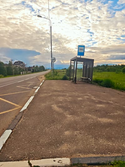 The image shows a roadside scene in Курово, Russia. A bus stop shelter with a blue bus stop sign is situated next to a paved area adjacent to a two-lane asphalt road. The road has yellow no-parking markings. A lamppost and a road sign are visible further down the road. In the background, there is a green field and forested area. There is a building set further back from the road. The sky is cloudy. There are no people visible in the image.