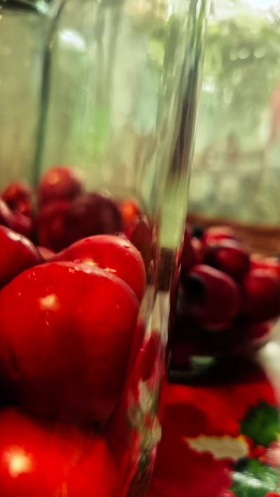 The image shows a close-up, shallow depth-of-field shot of bright red cherries inside a clear glass container. The cherries are glistening with small water droplets and some appear to have a dark stem. The background is blurred, showing hints of green foliage and a reddish-colored surface with a green, leafy pattern, possibly a tablecloth or placemat. The lighting creates highlights on the cherries, emphasizing their roundness and texture. The image is framed vertically. There is no visible indication of the city of Troițcoe, Moldova, within the visual content of the image.