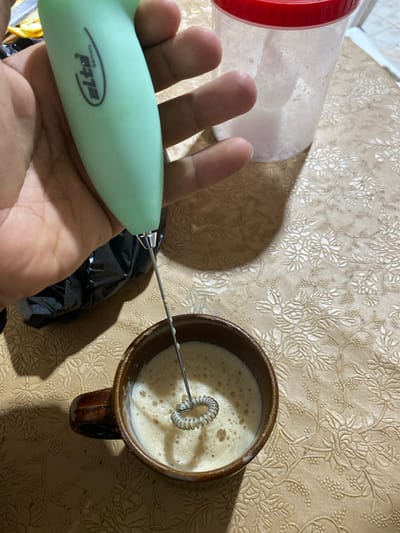 The image captures an indoor scene in Sidi Chahmi, Algeria, depicting a close-up view of a person's hand operating a milk frother. A right hand, exhibiting a medium skin tone, firmly grasps a light green "elta Germany" brand electric milk frother. The frother's coiled metallic whisk is partially submerged in a dark brown, stoneware-style ceramic mug, actively mixing a light brown liquid topped with a significant layer of white foam. To the right of the hand, a transparent plastic container with a red lid is visible, containing a white, foamy substance. A crumpled black object, possibly a plastic bag, is partially discernible behind the frother's handle. All objects are positioned on a light-colored, possibly beige or cream, tablecloth featuring a repeating embossed or patterned floral motif. Directional lighting from the right casts a distinct shadow of the hand and frother across the tablecloth.