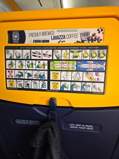The image shows a safety instruction card on a yellow surface, likely inside an airplane. The card features diagrams and text in multiple languages, including English, Polish, and what appears to be Chinese. The safety instructions cover topics such as fastening seat belts, using a life vest, and emergency exits. There is also an advertisement for "Freshly Brewed Lavazza Coffee" with a QR code for access to the menu. The yellow surface is part of an airplane seat. Below the safety card, on a dark blue surface, are two lines of text: "FASTEN SEAT BELT WHILE SEATED PROSZĘ ZAPIĄĆ PASY BEZPIECZEŃSTWA NIEZWŁOCZNIE PO ZAJĘCIU MIEJSCA" and "LIFE VEST IN PANEL ABOVE YOUR HEAD". A dark garment, possibly a jacket, is hanging from a dark blue hook attached to the blue surface. The location is described as Balice, Poland, which is the location of John Paul II International Airport Kraków–Balice. Therefore, the scene is inside an airplane at or near this airport.