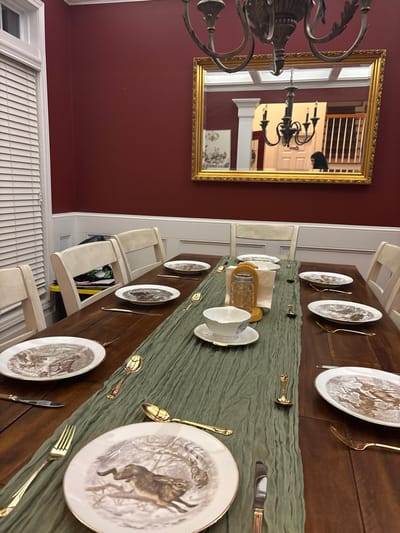 A long wooden dining table is set with plates depicting hares, gold-toned cutlery, and a sage green runner. Chairs surround the table, some with cushions. A gold-framed mirror hangs on the red wall to the left, reflecting a staircase and chandelier. A chandelier is visible hanging above the dining table. Window blinds cover the bottom of the wall.