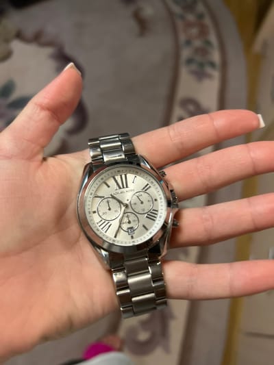 The scene is located in Dmitriyevskaya, Russia. A fair-skinned human hand is centrally positioned, displaying a silver-toned Michael Kors chronograph wristwatch. The watch features a circular white dial with black Roman numerals (II, IV, VI, VIII, X, XII) and stick hour markers. "MICHAEL KORS" is printed below the 12 o'clock position. Three circular subdials and a rectangular date window at the 6 o'clock position are visible on the dial. The watch case and metal link bracelet are reflective silver-toned. The hand is holding the watch face-up, with fingers slightly curled around the watchband and case. The background is an out-of-focus indoor environment, displaying a patterned surface, likely a carpet or upholstery, in muted beige and brown tones with blurred darker floral or abstract motifs.