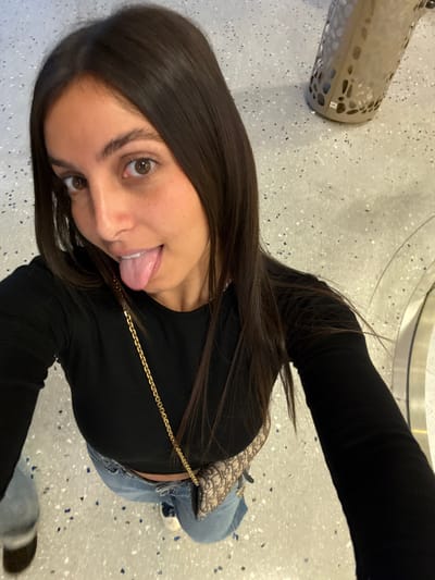 A female subject is depicted from a high-angle perspective, looking directly into the camera with her tongue extended. She possesses long, dark brown hair, brown eyes, and a fair complexion. She is dressed in a black long-sleeved crop top and light blue jeans. A beige and brown monogrammed handbag with a gold-colored chain strap is slung across her body. Partially visible footwear includes a dark shoe on the left and a light-colored sneaker on the right. The environment features a light-colored terrazzo-style floor speckled with white, grey, and dark blue flecks. In the upper right quadrant, a perforated, cylindrical beige object, possibly a waste receptacle or planter, bearing a "48" label, is present. A curved, reflective surface is visible in the lower right background. The scene is set indoors within a public space in Saint-Laurent-du-Var, France.