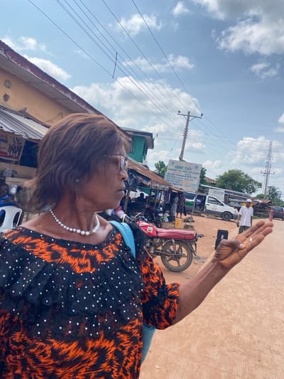 In Amaseri, Nigeria, a woman in a black and orange patterned top and a pearl necklace stands outdoors in front of a row of stalls. Her right arm is extended, palm open, in a gesturing motion. Behind her, a red motorcycle is parked. In the background, a white van and other vehicles are visible, along with a billboard displaying "EBONYI STATE COMMUNITY AND SOCIAL DEVELOPMENT". Power lines crisscross the sky, and scattered clouds drift overhead. The ground is a reddish-brown dirt road.