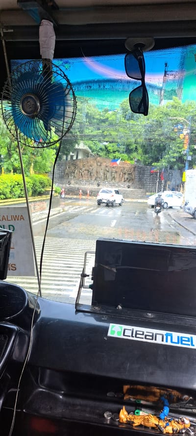 This image captures a view from inside a vehicle, likely a jeepney or a similar public transport in the Philippines. The perspective is from the driver's or front passenger's seat, looking out through the windshield. The windshield is wet, suggesting it is raining or has recently rained.

Prominent objects in the foreground include a blue electric fan mounted to the left side of the windshield, its cord wrapped with white tape. A pair of dark-rimmed sunglasses is attached to the windshield by a suction cup. A sticker with the text "cleanfuel" and a green recycling symbol is visible on a black dashboard component on the right. There are also a couple of small action figures, one gold and orange with a blue accessory and another in gold, positioned on the dashboard.

The scene outside the windshield shows a street with vehicles, including a white car and a motorcycle. There is a raised stone structure with bas-relief carvings, possibly a monument, adorned with a Philippine flag. Lush green trees and buildings are visible in the background. The road appears wet, reflecting the surroundings.

The overall impression is a snapshot of daily life in a busy urban environment during inclement weather. The presence of the fan and sunglasses suggests attempts to manage comfort within the vehicle despite the conditions. The visible text "cleanfuel" and the recycling symbol hint at a focus on environmental consciousness or a brand affiliation.