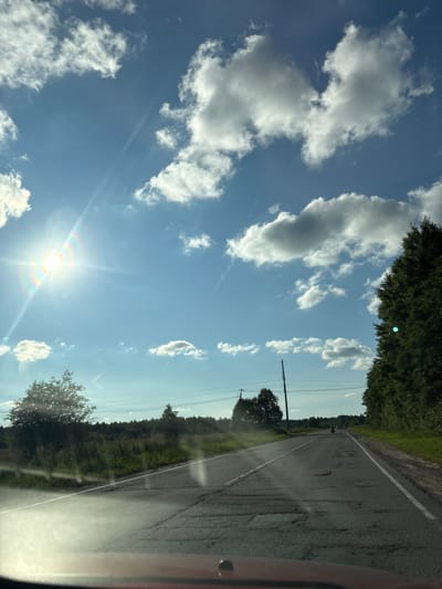 A bright sun shines through a blue sky dotted with white clouds, casting a rainbow glare on the left side of the image. The sun's rays streak across the sky. Below the sky, a two-lane asphalt road stretches into the distance, its surface cracked and uneven with numerous potholes. A white line marks the left edge of the road, and a broken white line divides the two lanes.
On the left side of the road, tall green grass and scattered trees line the shoulder. Further back, a dense forest stretches across the horizon. On the right side of the road, a line of tall green trees creates a natural border. In the distance, a motorcycle with two riders is visible on the road, heading away from the camera. Two utility poles stand along the right side of the road in the mid-ground. The foreground shows the dashboard of a vehicle, implying the photo was taken from a car. The environment appears to be rural, consistent with the location in Zamost'e, Russia.
