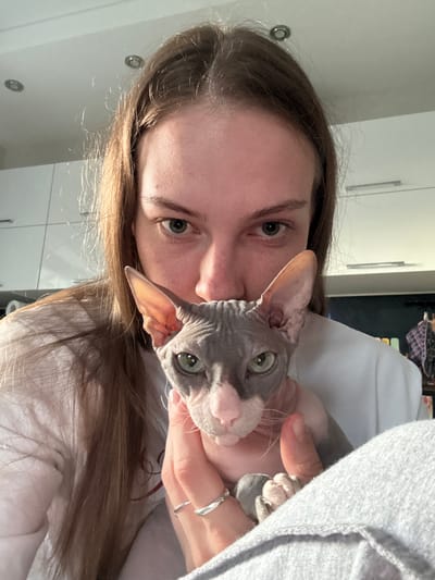 The image shows a close-up of a person holding a hairless cat, likely a Sphynx, in what appears to be an indoor setting. The person has long brown hair and is looking directly at the camera with green eyes. The cat has gray and white fur with large ears and green eyes. The background suggests a domestic environment with white cabinets and what look like ceiling lights. The person is wearing a white t-shirt and the cat is being held in their hands, with the cat's paws visible. The overall impression is of a casual, personal moment captured by a selfie. There is no indication of the location being Novaya Ivanovka, Russia.