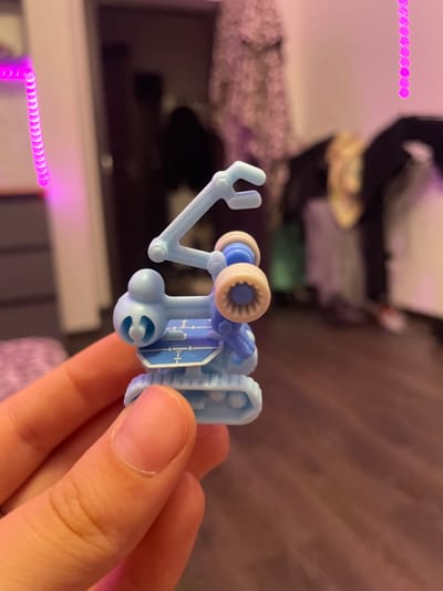 A close-up shot shows a small, light blue toy robot being held by a person's hand. The robot is intricate, with a mechanical arm, treads, and a central body featuring blue accents and what appear to be control panels or decorative elements. The background is blurred, suggesting an indoor setting with soft lighting. Hints of pink and purple light, possibly from LED strips, are visible on the left and right edges of the image, creating a diffused glow. The floor appears to be made of dark wood planks. The overall scene is focused on the toy, with the surroundings serving as a soft, atmospheric backdrop. There is no information in the image that can confirm or deny the location of Borisova Griva, Russia.