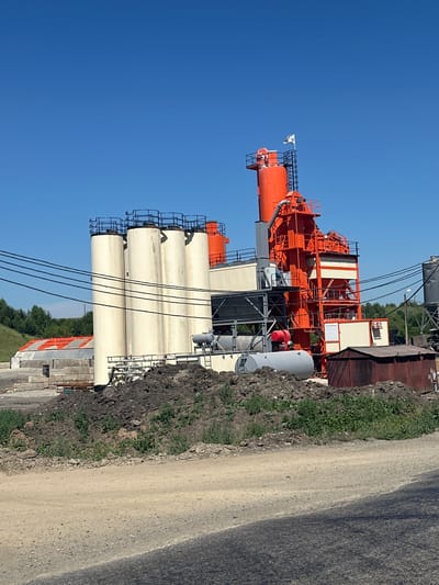 An asphalt mixing plant is visible on a clear, sunny day in Nizhnekamenka, Russia. The plant consists of several large, cylindrical storage silos, painted in a light cream color, standing vertically. Adjacent to these silos is a tall, complex structure, primarily painted in bright orange, with multiple levels, platforms, and connecting pipes and chutes. A white flag with a logo is flying from the top of the orange structure. Several large, dark wires run horizontally across the mid-section of the image, obscuring parts of the plant. In the foreground, there is a large pile of dark soil or dirt, with some green vegetation growing on it. Behind the silos, a long, low building with an orange, corrugated roof is partially visible. To the right of the main plant, a rusty, rectangular metal shed or container sits on the ground, partially obscured by the pile of dirt. The ground in the foreground is a dusty, gravelly road. In the background, rolling green hills covered with trees extend to the horizon under a clear blue sky. No people are visible in the image.