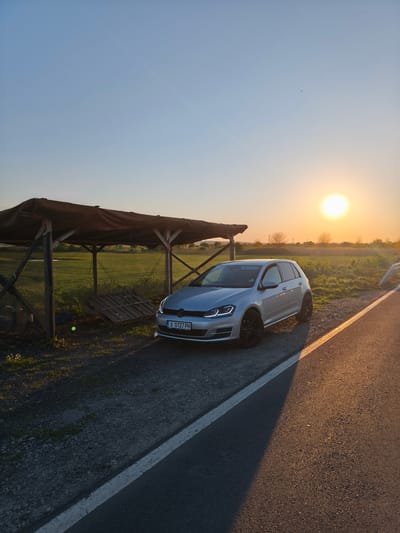 The image shows a silver Volkswagen Golf parked near a small, wooden shelter with a tarp roof next to a road. The car has a Bulgarian license plate "A 0127 PH." The sun is setting in the background, casting a warm glow over the scene. The location appears to be rural, with a field visible behind the shelter. The road has a white dividing line. This is in the Trastikovo region of Bulgaria.