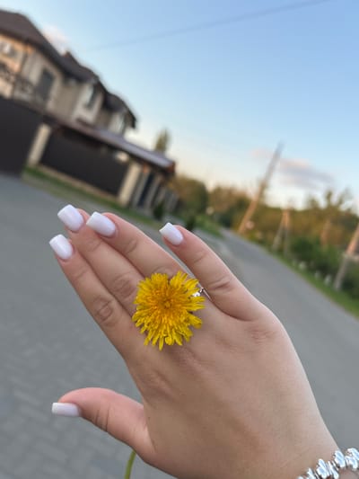 A close-up shot depicts a fair-skinned hand holding a yellow dandelion. The hand is adorned with square-shaped white acrylic nails and a silver bracelet is visible on the wrist. A silver ring is worn on the middle finger. The background shows a residential street with modern-looking houses, trees, and a gray asphalt road. The location appears to be Chernomurovskii, Russia.