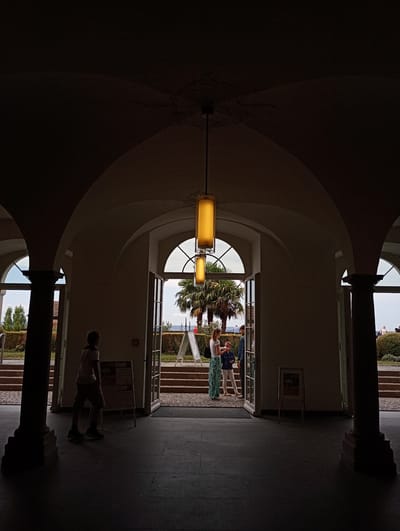 The image depicts an interior view of a building with arched ceilings and columns, looking out towards an exterior landscape. In the foreground, a young person, silhouetted against the light, walks away from the viewer towards the left. They are wearing a t-shirt and shorts. Further back, a woman in a long, patterned skirt and a light-colored top, and a child wearing a blue jacket and white pants, are standing near an open doorway. They appear to be interacting. Beyond them, a palm tree is visible, with stairs leading up to a garden area with more trees and shrubs. The exterior also shows a body of water, likely Lake Constance, and distant hills. There are several A-frame signs visible both inside and outside the building. The lighting suggests it is daytime, with strong backlighting from the exterior creating silhouettes of the interior architecture and people. This scene is located at the entrance to the Meersburg Castle (Alte Schloss Meersburg) in Meersburg, Germany.