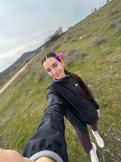 A selfie-style photograph captures a single adult female standing on a grassy hillside under an overcast sky in the vicinity of Ostar Kamak, Bulgaria. The female, with long dark brown hair pulled into a ponytail, is wearing a black long-sleeved athletic jacket with a white Nike logo on the upper left chest, maroon leggings, and light-colored athletic shoes with white socks. Two small purple flowers are placed in her hair above her left ear. Her left arm is extended towards the camera, holding the recording device. She looks directly forward with a faint smile. The immediate environment consists of green grass interspersed with patches of dry vegetation and sparse, leafless shrubs on a sloping terrain. In the background, a larger, forested hill or mountain range is visible under a uniformly grey sky.
